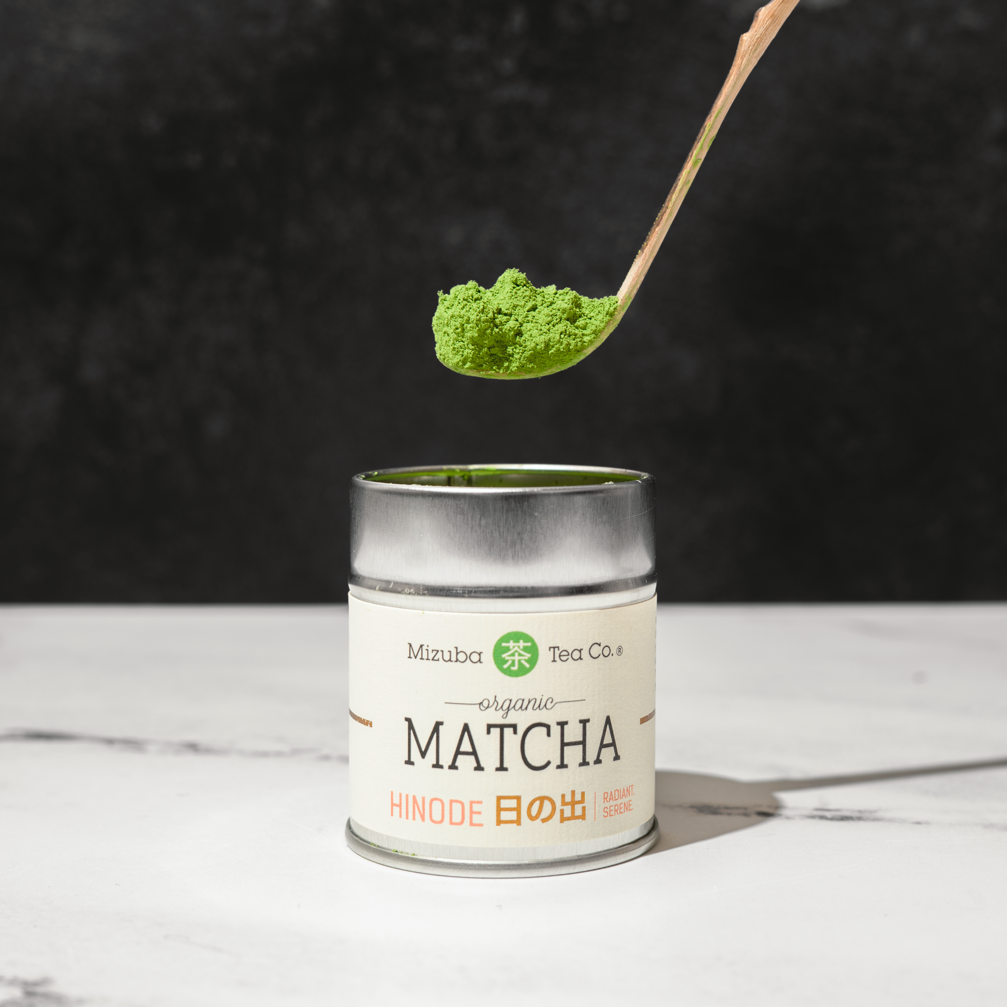 Can of Mizuba Matcha with a chashaku spoonful of green powder above it on a dark background. 