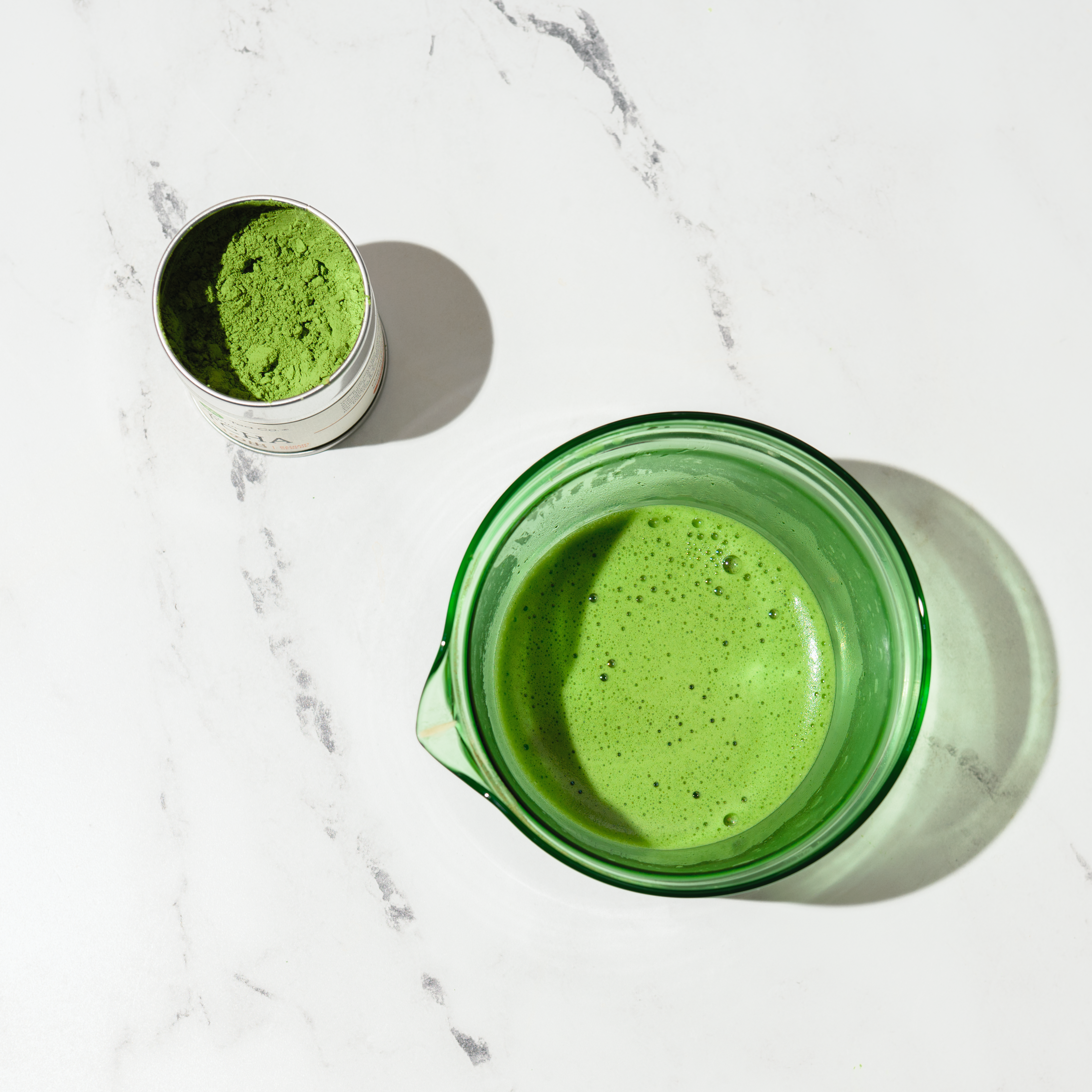 Frothy matcha green tea in a glass bowl with a container of Mizuba matcha powder on a marble surface.