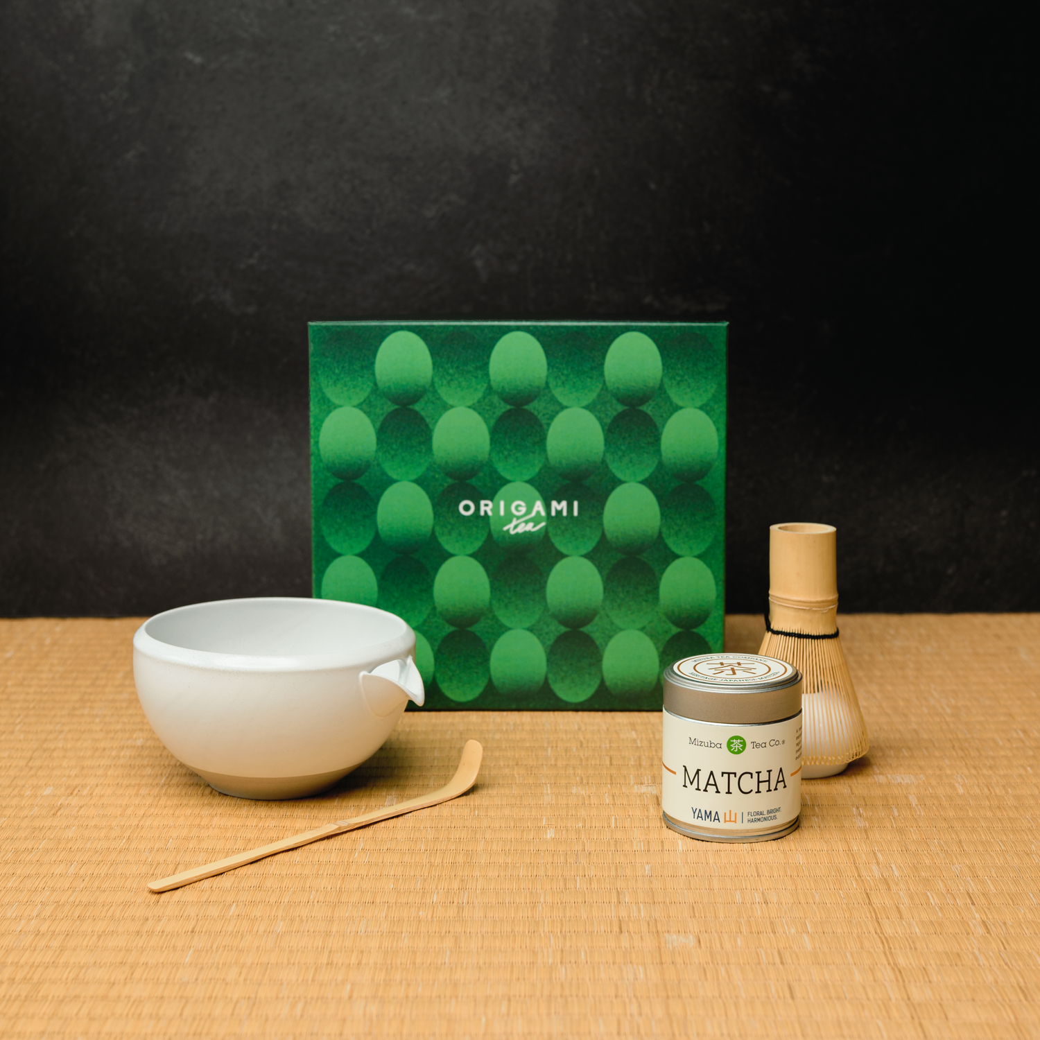 Matcha-making set with a matcha chawan tea bowl, whisk, and Yama Mizuba matcha canister on a tatami mat surface with a black background.