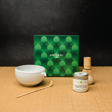 Mizuba Origami Matcha tea set featuring Shirakawa Matcha with a bowl, whisk, and container on a tatami mat surface with a green Origami box in the background.