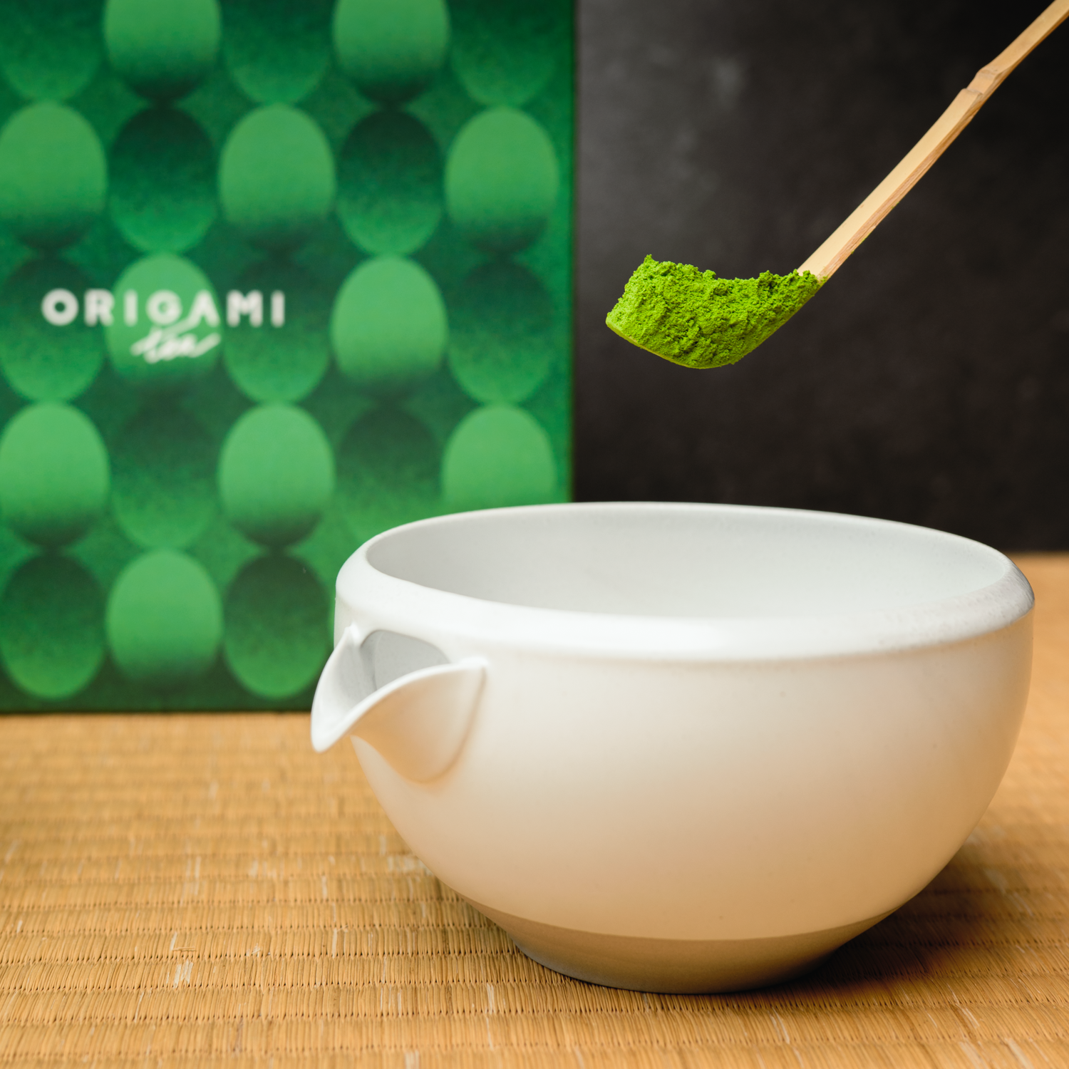 White ceramic matcha bowl with a wooden spoon holding green matcha powder, Origami Tea box in the background.