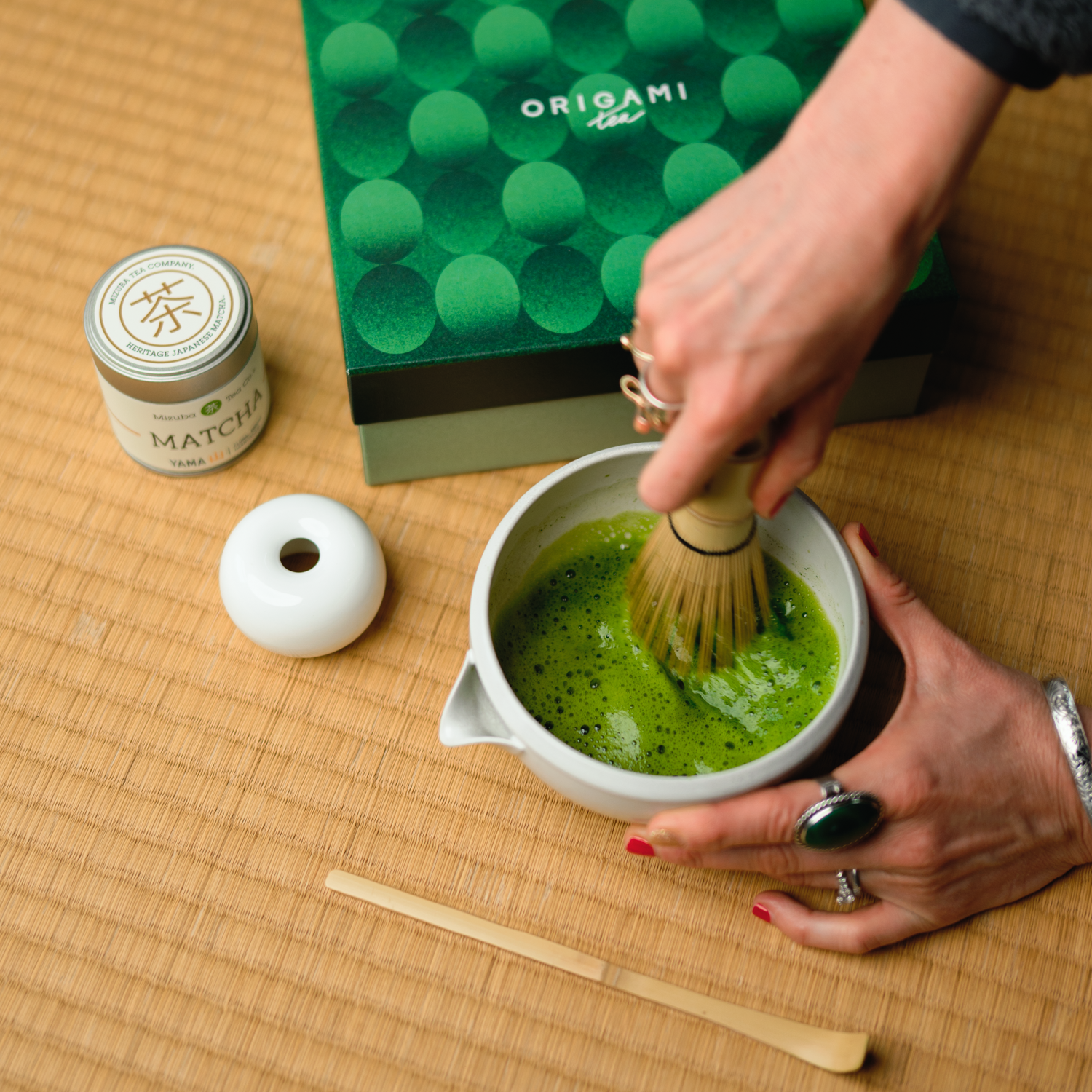 Person preparing Mizuba Yama matcha tea with a bamboo whisk on a tatami mat, next to an Origami matcha gift box.