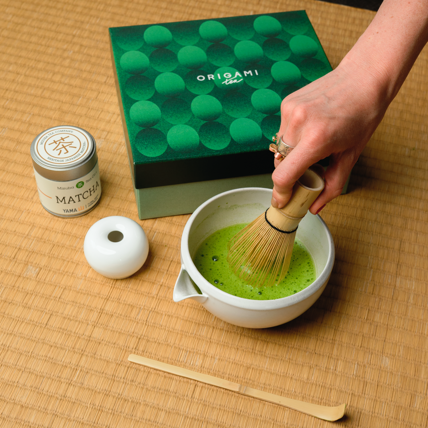 Person preparing Mizuba Yama matcha tea with a whisk on a tatami straw mat, next to an Origami gift box and matcha container.