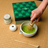Person preparing Mizuba Yama matcha tea with a whisk on a tatami straw mat, next to an Origami gift box and matcha container.