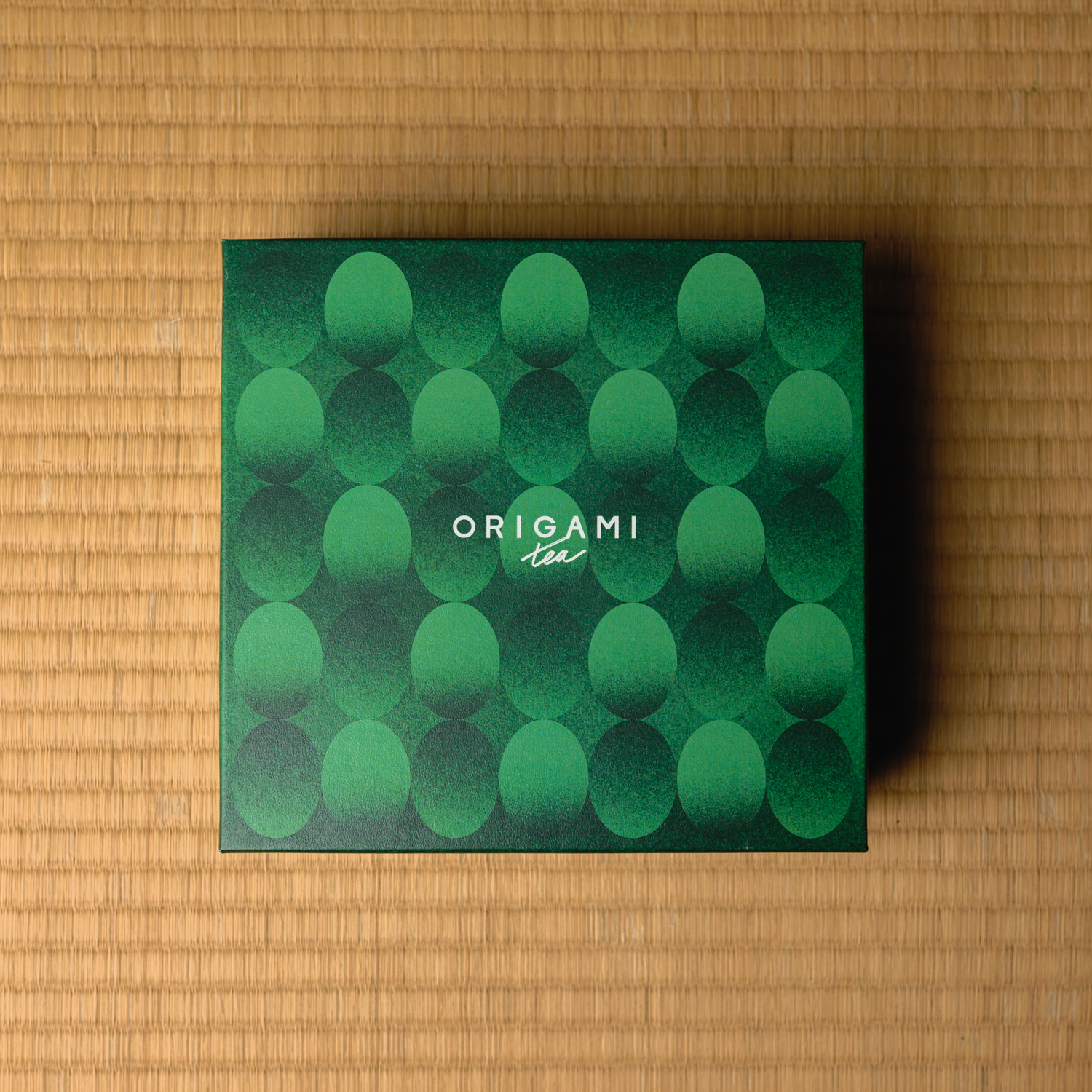 Green box with circular pattern and 'Origami Tea' branding on a tatami straw surface.
