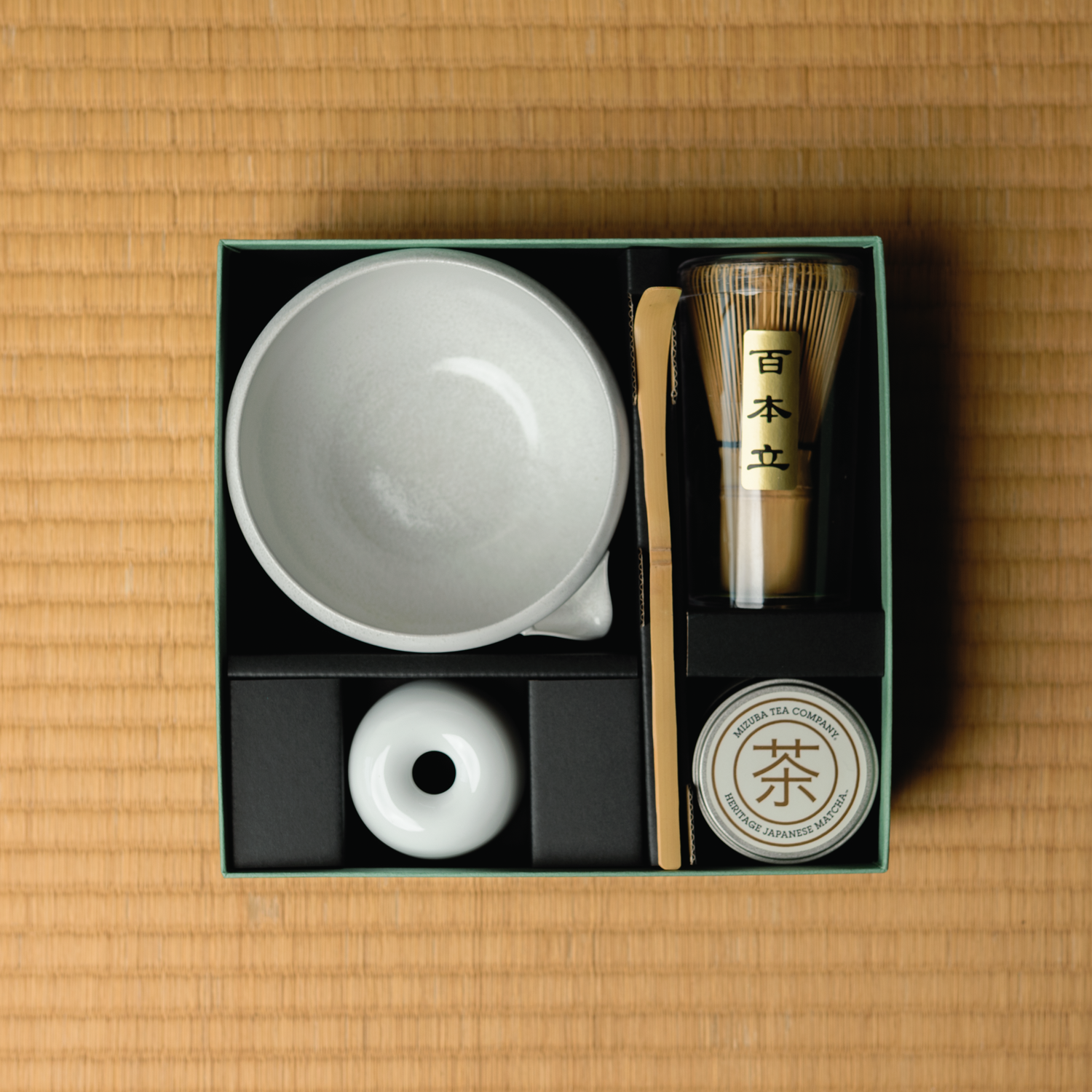 Mizuba Origami matcha tea set with a white chawan tea bowl, whisk stand, bamboo matcha whisk, tin of matcha green tea, and chashaku tea scoop, and accessories in a box on a tatami surface.