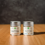 Two tins (Hinode and Yorokobi) of Mizuba Tea Co. Matcha green tea on a wooden surface with a dark background.