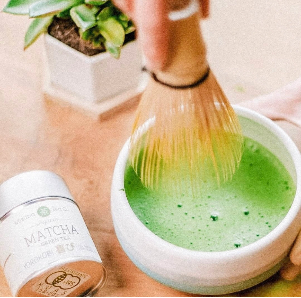 A tin of Mizuba Yorokobi Matcha Green Tea lies next to a cup of freshly whisked matcha. A chasen bamboo whisk hovers above. 