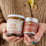 A person in a chunky, beige, knit cardigan holds two jars of gourmet food products: a jar of Miso Caramel by Sugarpine Drive In and a tin of Hojicha from Mizuba Tea Co.