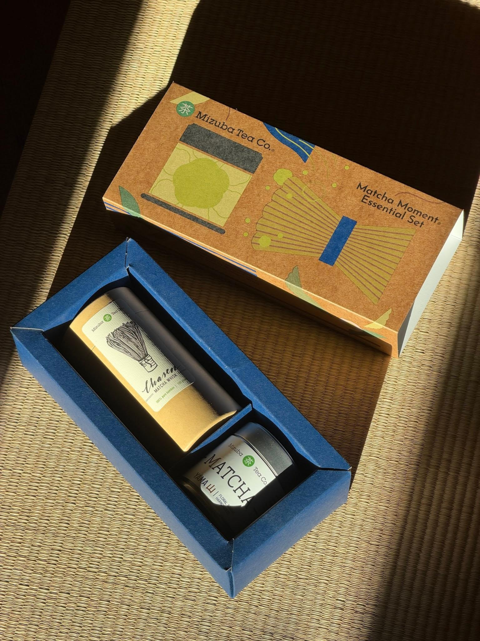 A boxed , Mizuba Tea Co. tea gift set displayed on a woven tatami mat, with the open blue tray containing a matcha tin and a chasen whisk alongside the illustrated outer box labeled 'Masaba Tea Co.' and 'Matcha Moment Essential Set.