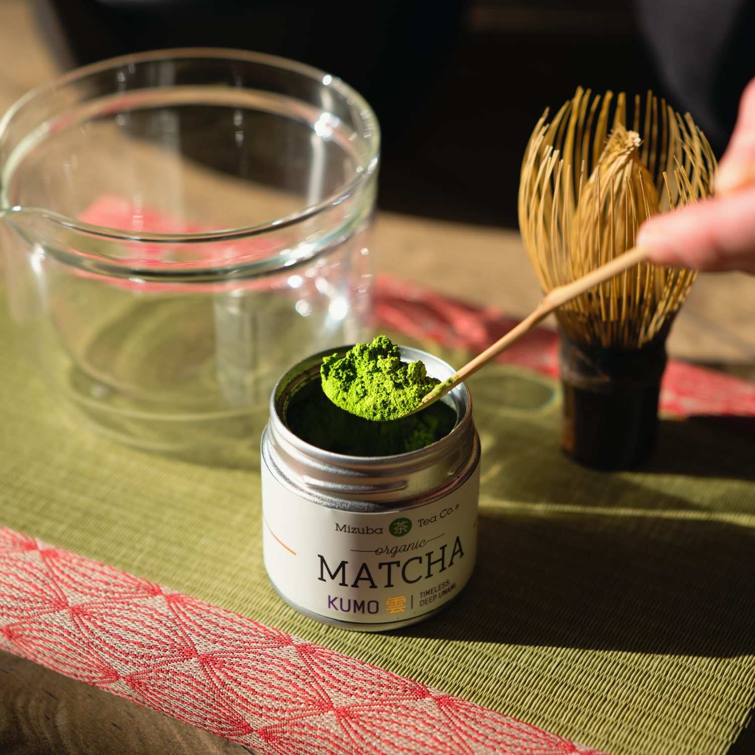 A person lifts verdant, rich organic Japanese matcha tea with a chasahku scoop above the tin. There is a chasen whisk and a glass chawan behind it on a tatami mat. 