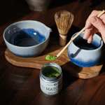 Person preparing matcha tea with a container of matcha powder on a wooden table. A blue and white chawan, bamboo chasen, and blue ceramic cup are also on the table.