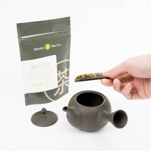 A 50g bag of organic Genmaicha tea by Mizuba Tea Co. stands to the left of a black clay tea pot. A hand is putting loose leaf tea into the pot from a cherrywood scoop.