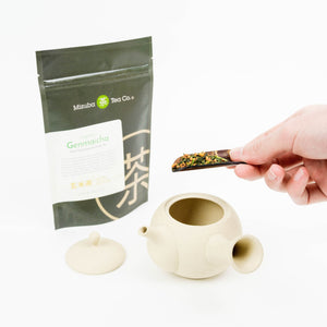 A 50g bag of organic genmaicha tea stands next to a white kyusu tea pot. A hand puts tea into the pot.